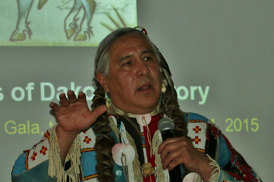 Head & shoulders of Monte Yellowbird dressed in native garb, talking into microphone w/ native slide on wall in background