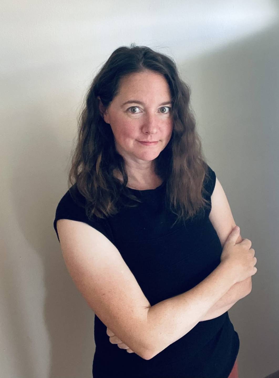 Ali Larock stands side facing arms crossed. Ali is wearing a black tee-shirt and has her long brown wavy hair down. She smiles slighlty as she look directly into the camera