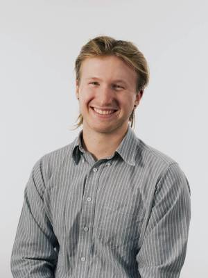 head and shoulders of Tobias Zikmund with short, dirty blonde hair, black dot earrings, pale skin and a big toothy smile; wearing a grey collared shirt with white stripes