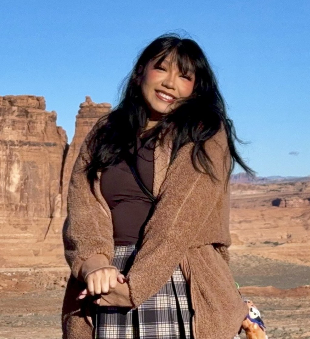 Native American Kiera Fox with a toothy smile and long black hair, standing in front of canyons on a sunny, blue sky day, wearing a light brown coat with a dark brown undershirt