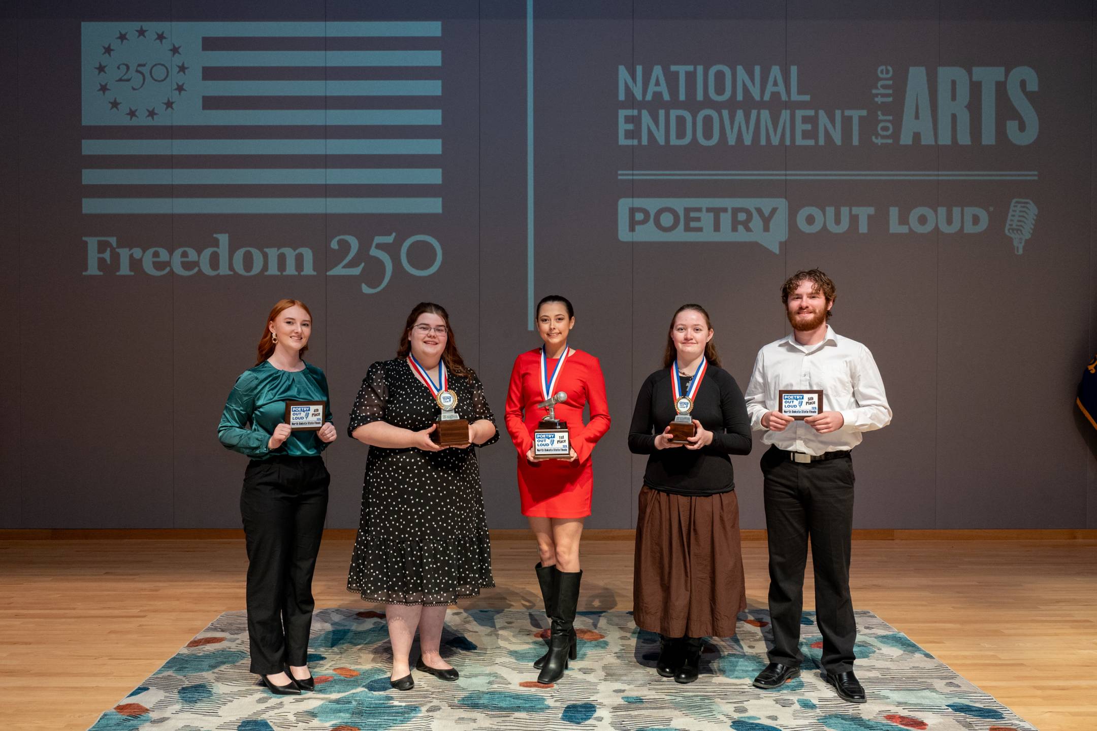 5 high school students standing on a stage, holding 1st-5th trophies received after participating in the 2026 ND Poetry Out Loud Competition in Bismarck