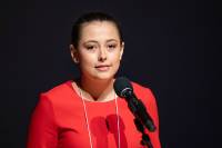 Head and shoulders of Wyndmere High School student Aspen Geist, wearing a red top, dark haired pulled back, standing in front of a microphone with a slight closed smile