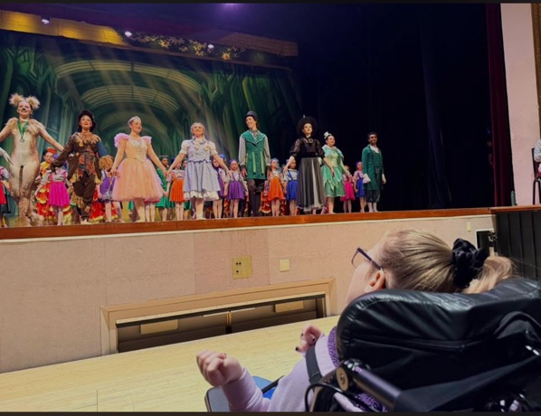 The cast of the Wizard of Oz on stage, holding hands, with a girl in a wheelchair looking on from below