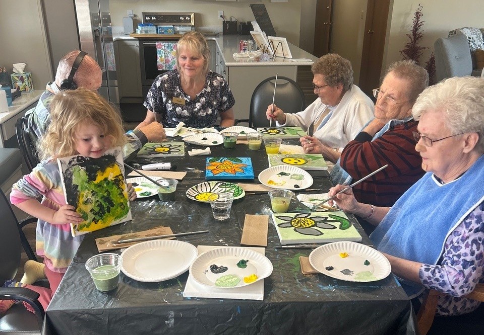 A young child and seniors sit around a table painting flowers on small canvases during an art activity.