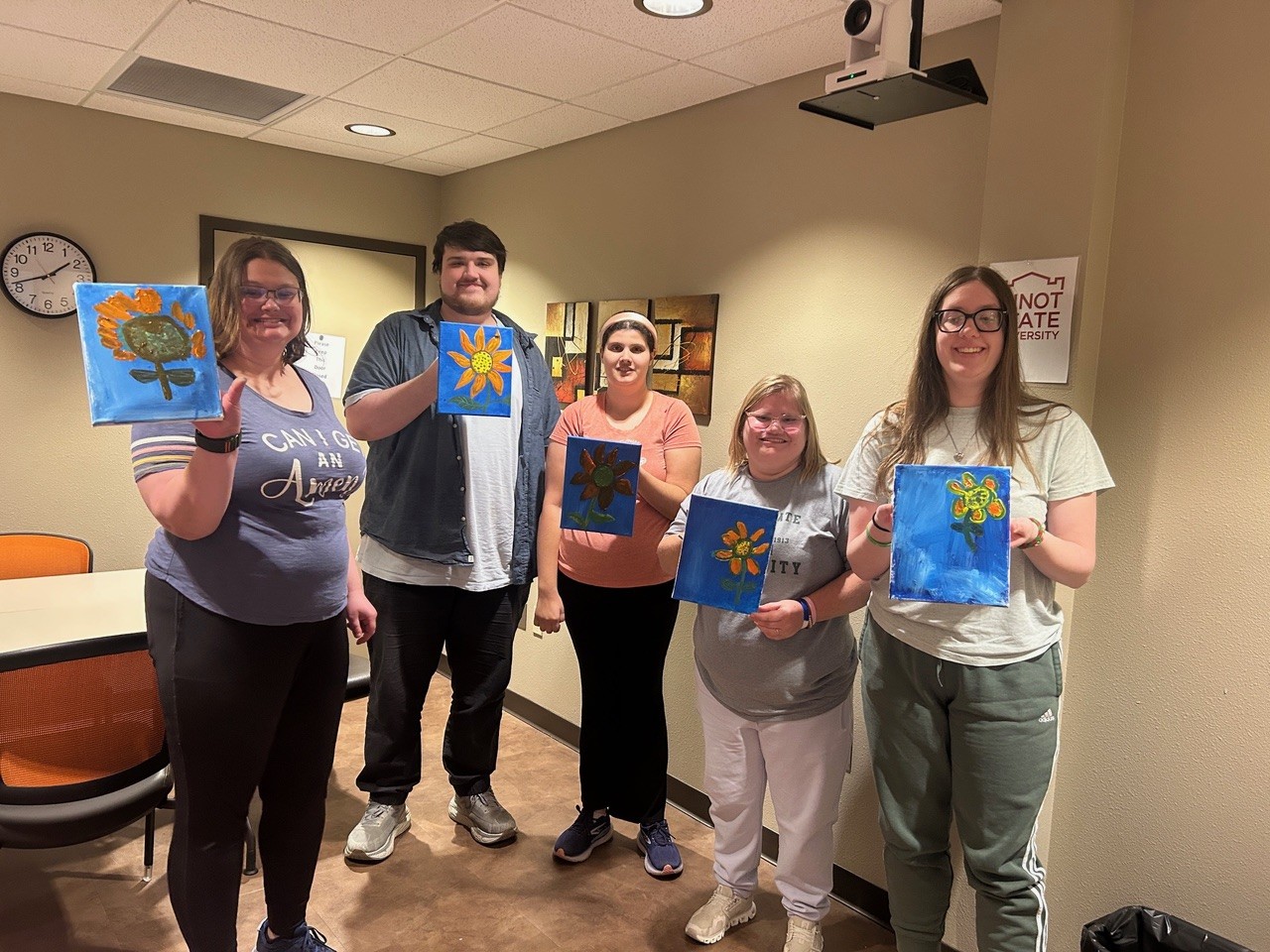 Five smiling people stand in a room, each holding up a small blue canvas painted with an orange and yellow flower