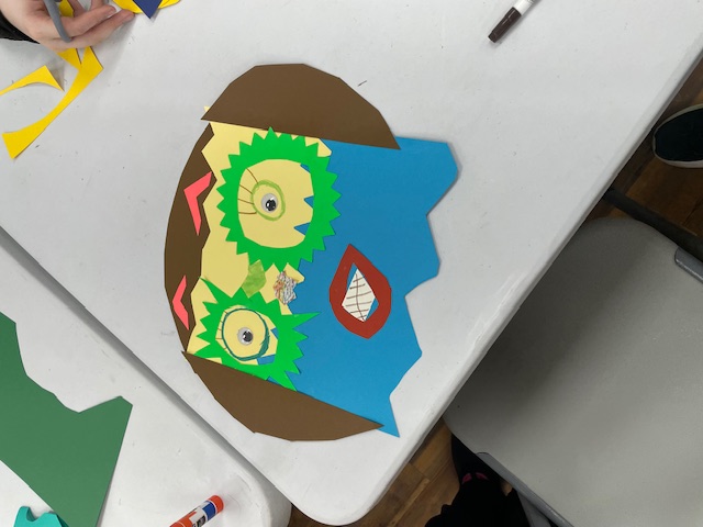 an abstract self-portrait made out of construction paper, showing green cut-out glasses, a mouth out-lined in red, and orange triangle eyebrows all on a square face
