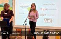NDCA August 2026 E-NEWS header showing a teaching artist helping a young elementary student in Kindred to speak into a microphone