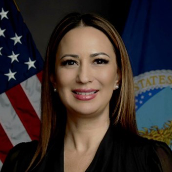 Head and shoulders of Kristy Zaste with light brown, long hair, dark eyes, slightly toothy smile with red lipstick; wearing a black v-neck shirt; with the American and North Dakota flags behind her