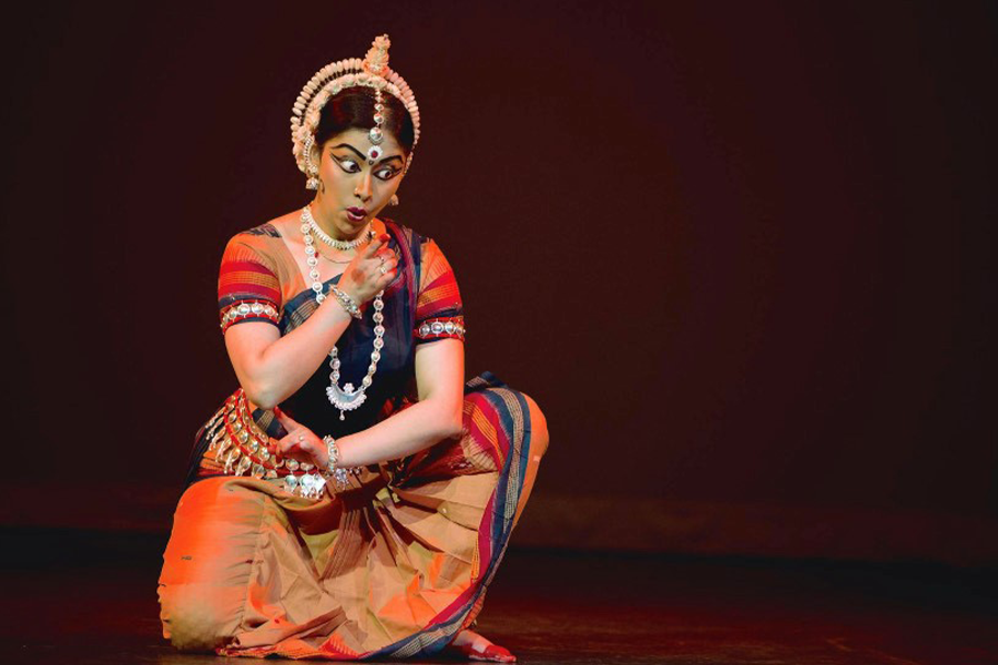 a woman wearing traditional Indian dance clothing and make-up, posing in a kneeling-style position with her arms rotating