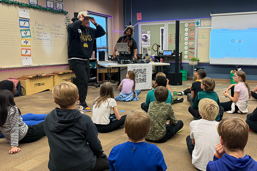 a classroom of elementary students sitting on the floor, looking up at a hip-hop artist/teacher who is performing with a man standing next to him, running the music with a computer and other sound equipment