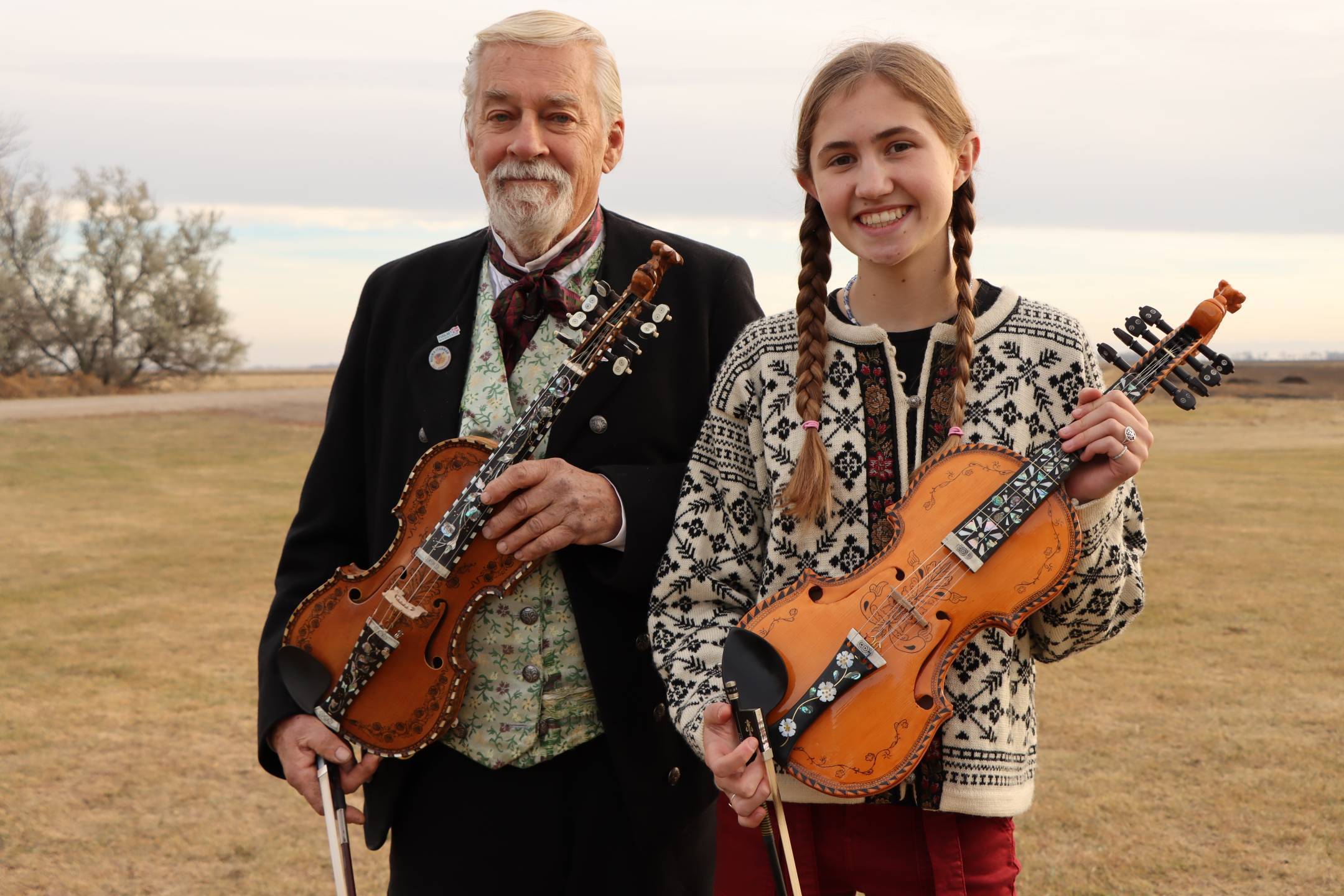 older man standing next to a young girl with braided hair, dressed in Norweigan clothing, both holding a hardanger fiddle, standing outside on a fall day