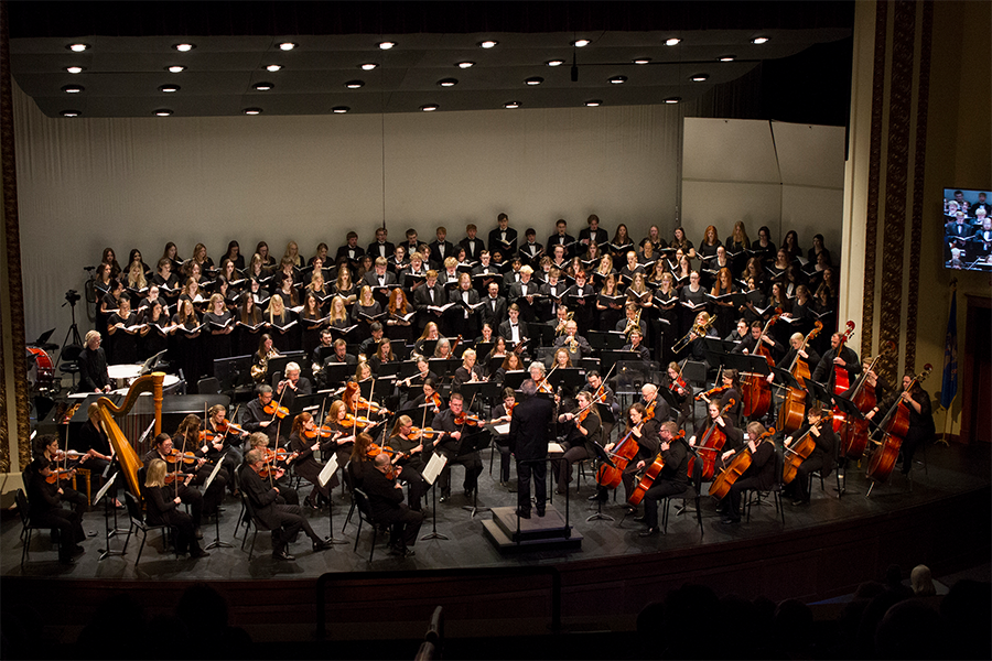 Minot Symphony orchestra playing on stage with a full concert choir singing behind them and a director in front; everyone is wearing black