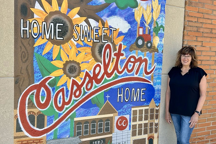 outdoor mural saying Home Sweet Casselton Home with sunflowers, buildings, tractors, and the female, middle-aged artist standing next to it