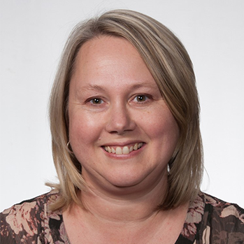 Head and shoulders of Amy Tichy, short dirty blonde, straight hair, pale skin, dark shirt with pale pink flowers, off shite background