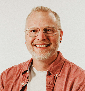 Head and shoulders of Matthew Anderson wearing a dark coral button-up shirt with a white undershirt, clear rimmed glasses, with pale skin, a big toothy smile, dark eyes, short, light hair and short gray beard
