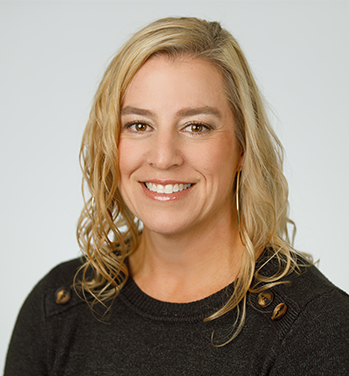 Head and shoulders of Rhea Beto with brown eyes, curly, shoulder-length blond hair, light skin, toothy smile, wearing dark long-sleeve shirt with light grey background