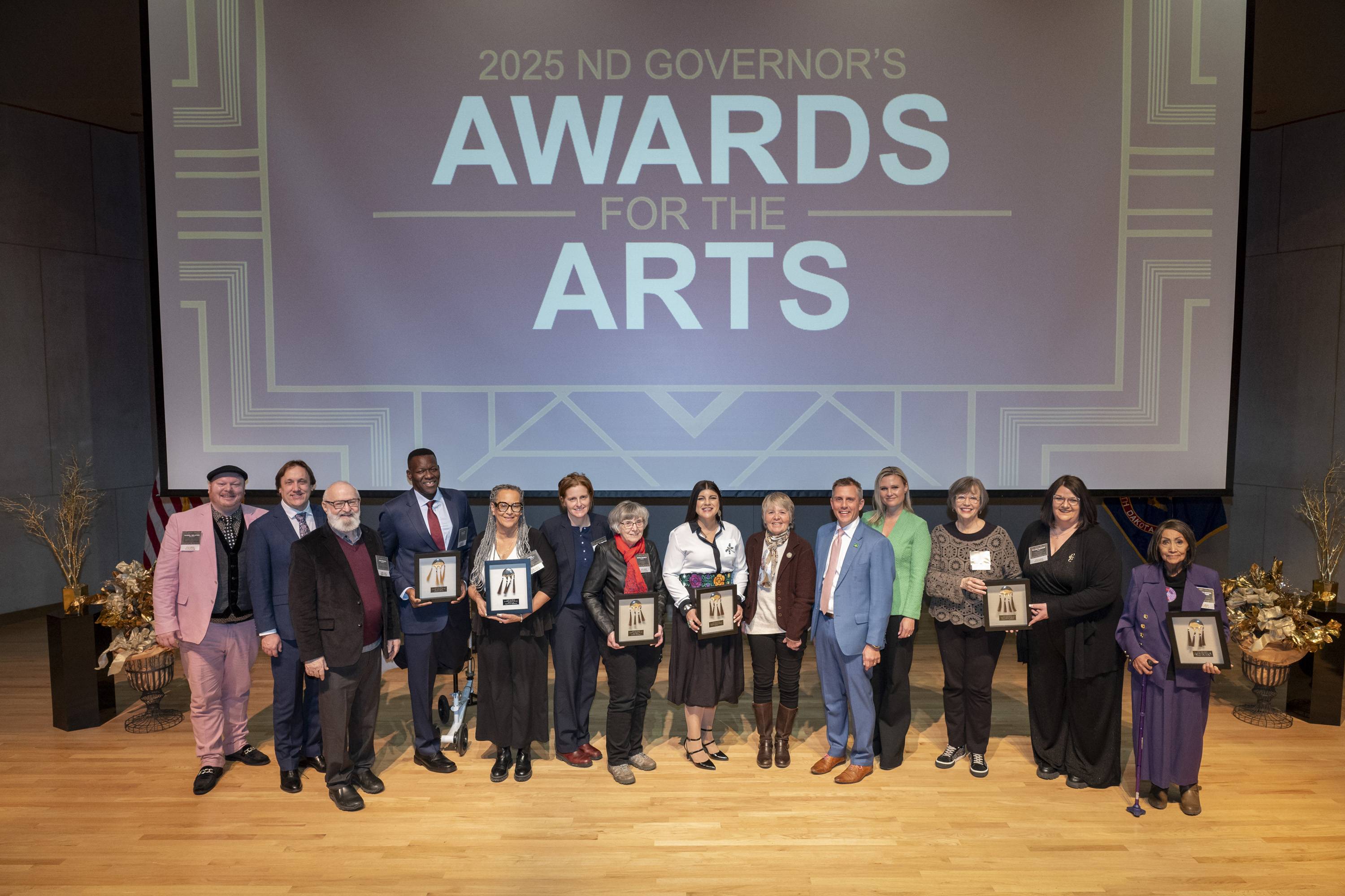 Large group photo with ND Governor's Awards for the Arts on screen above a stage with all seven recipients and ND Governor and First Lady Armstrong
