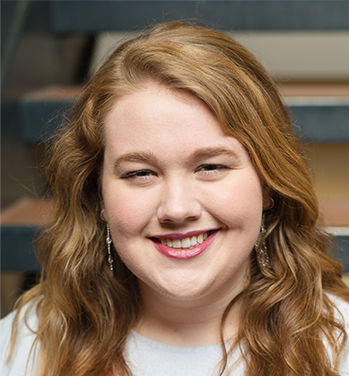 head and shoulders of Brenna Lahren with long, curly, red-brown hair, light skin, toothy smile with red lipstick, long dangling earrings, light grey shirt with blurred stairwell in the background