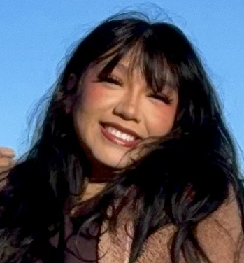 head and shoulders of Kiera Fox with long, straight, black hair, toothy smile with dark red lipstick, light brown jacket with dark brown under shirt and a blue sky and canyon background