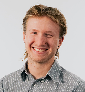 head and shoulders of Tobias Zikmund with short, dirty blonde hair, black dot earrings, pale skin and a big toothy smile; wearing a grey collared shirt with white stripes
