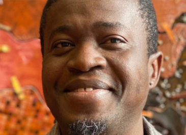 Hamzat Amoussa headshot with slight goatee, tightly shaved hair and a secretive grin