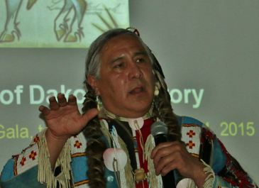 Head and shoulders of Monte Yellowbird in native apparel, talking on microphone w/ native horse image on slide projected behind him