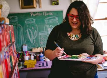 Teaching artist Nicole Gagner mixing paint, wearing red-rimmed glasses, with long, thick wavy dark hair, enjoying herself