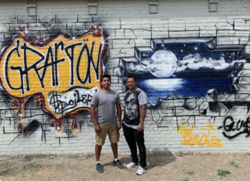 2 people standing in front of brick wall grafiti-painted that says Grafton