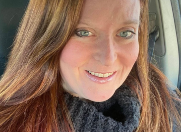 close up face and shoulders of artist Myra Klein with long, straight auburn hair, blue eyes, heavy grey turtle-neck sweater, pale skin, and slightly toothy smile