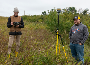 Two men standing outside on an overgrown field on a fall day with surveying equipment