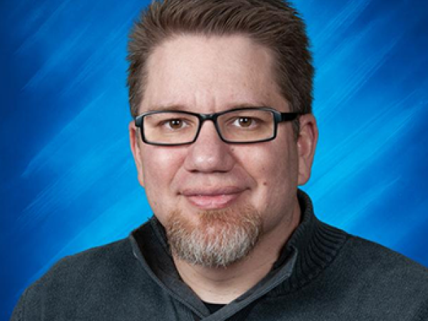 Formal chest-up image of Derek Morin wearing a collared dark-grey sweater with a full-head of dark hair, salt and pepper goatee, dark-rimmed rectangular glasses, light skin and a closed smile