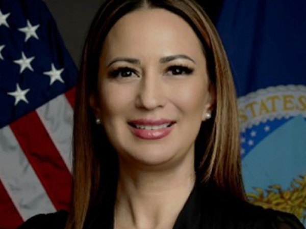 Head and shoulders of Kristy Zaste with light brown, long hair, dark eyes, slightly toothy smile with red lipstick; wearing a black v-neck shirt; with the American and North Dakota flags behind her