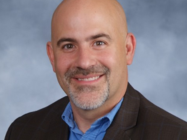 Head and shoulders of Shawn Oban, bald, pale skin, goatee, dark suit jacket with blue button-up shirt on blue cloudy background