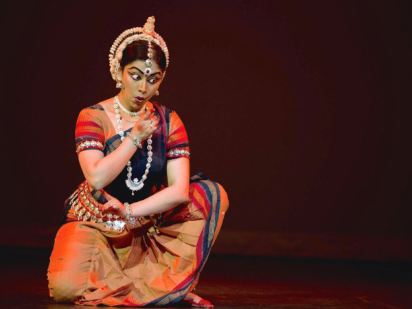 a woman wearing traditional Indian dance clothing and make-up, posing in a kneeling-style position with her arms rotating