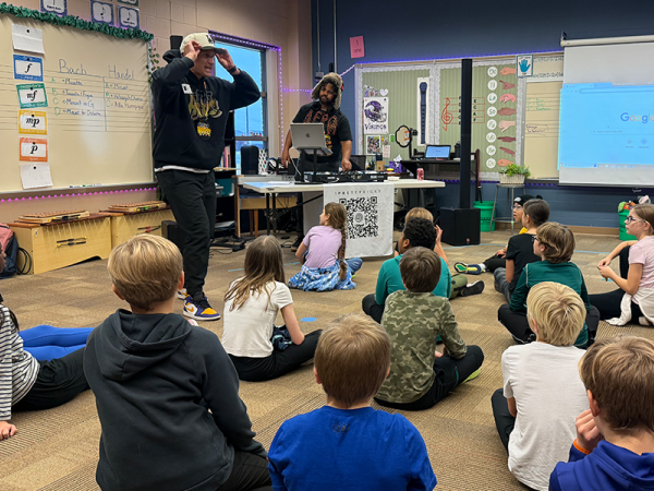 a classroom of elementary students sitting on the floor, looking up at a hip-hop artist/teacher who is performing with a man standing next to him, running the music with a computer and other sound equipment