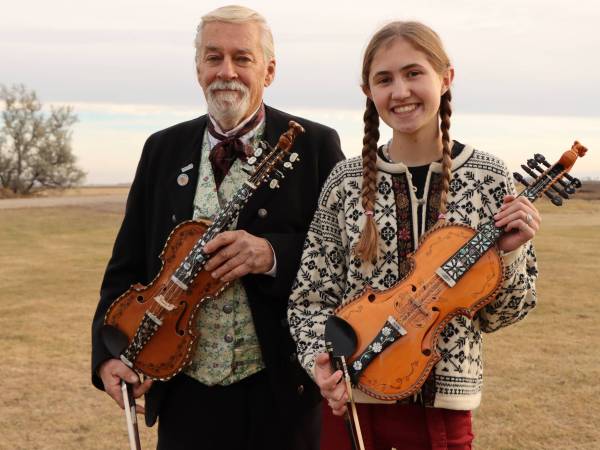 older man standing next to a young girl with braided hair, dressed in Norweigan clothing, both holding a hardanger fiddle, standing outside on a fall day