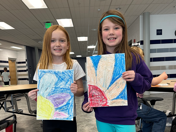 Two elementary-aged girls, smiling and holding artwork they each created that shows the side of a sun-flower