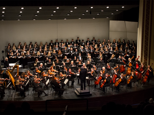 Minot Symphony orchestra playing on stage with a full concert choir singing behind them and a director in front; everyone is wearing black