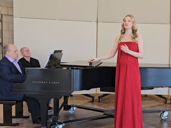 a woman standing on stage, wearing a formal, strapless, long red dress, singing beside a black concert piano with two people playing it