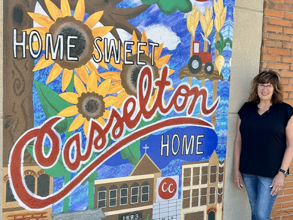 outdoor mural saying Home Sweet Casselton Home with sunflowers, buildings, tractors, and the female, middle-aged artist standing next to it