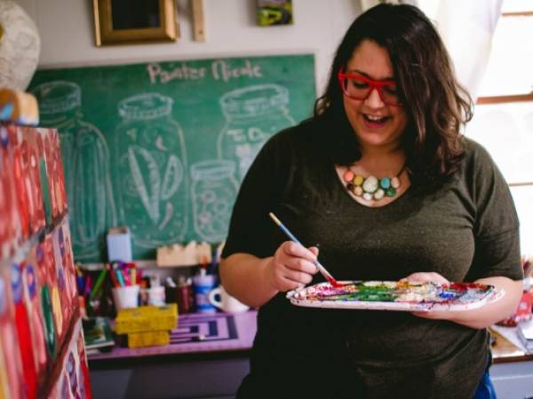 Teaching artist Nicole Gagner mixing paint, wearing red-rimmed glasses, with long, thick wavy dark hair, enjoying herself