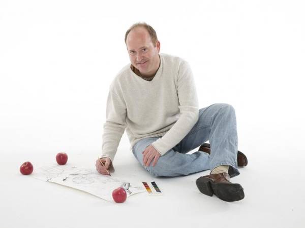 Teaching artist Markus Tracy with balding brown hair, smiling brightly, sitting on a white floor with crossed legs, wearing blue jeans and a white sweater, playing a game with red balls