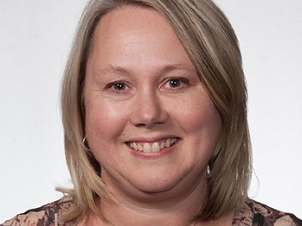Head and shoulders of Amy Tichy, short dirty blonde, straight hair, pale skin, dark shirt with pale pink flowers, off shite background