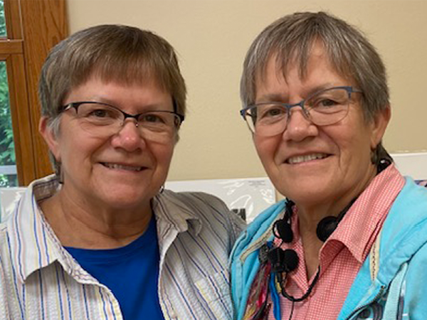Head and shoulders of sisters Barbara and Beverly Benda, with short brown/grey hair, matching cat-style glasses and toothy smiles