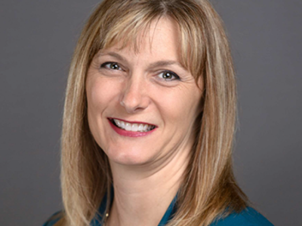 Head and shoulders of straight, blonde-haired, brown-eyed Christi Stonecipher on grey background wearing teal jacket top