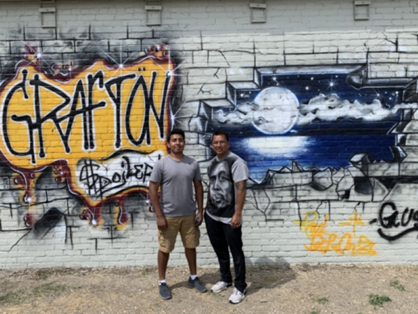 2 people standing in front of brick wall turned graffiti mural with Grafton spelled out