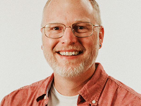 Head and shoulders of Matthew Anderson wearing a dark coral button-up shirt with a white undershirt, clear rimmed glasses, with pale skin, a big toothy smile, dark eyes, short, light hair and short gray beard