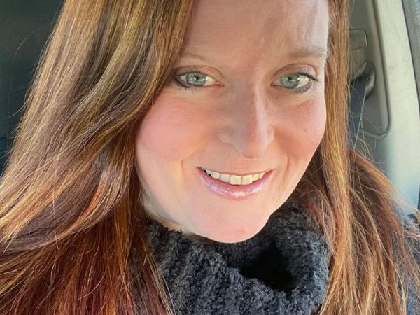 close up face and shoulders of artist Myra Klein with long, straight auburn hair, blue eyes, heavy grey turtle-neck sweater, pale skin, and slightly toothy smile
