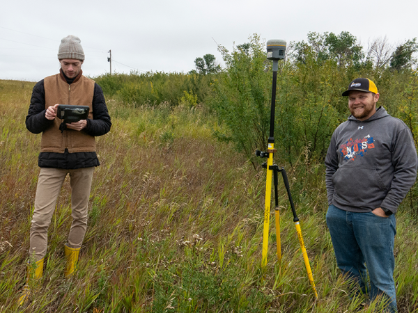 Two men standing outside on an overgrown field on a fall day with surveying equipment