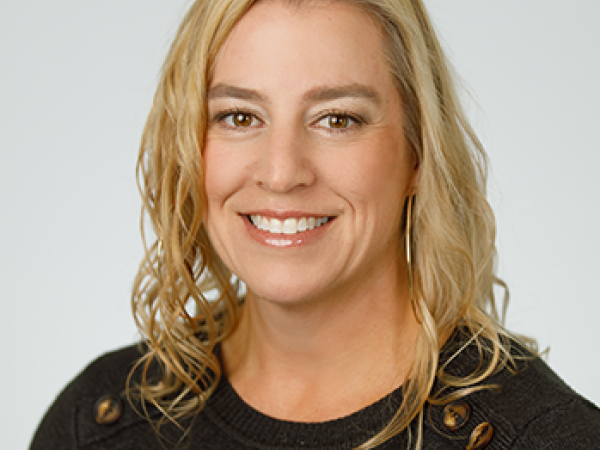 Head and shoulders of Rhea Beto with brown eyes, curly, shoulder-length blond hair, light skin, toothy smile, wearing dark long-sleeve shirt with light grey background